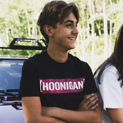 Young man wearing a Hoonigan x Lia Block STAR CENSOR BAR Short Sleeve T-Shirt, smiling with arms crossed beside a car.