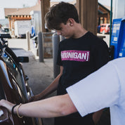 Young man wearing a black Hoonigan x Lia Block STAR CENSOR BAR T-shirt at a gas station, focused on a car.
