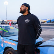 Man wearing a black Hoonigan BRACKET X LOGO long sleeve T-shirt, standing beside a damaged car at a motorsport event.