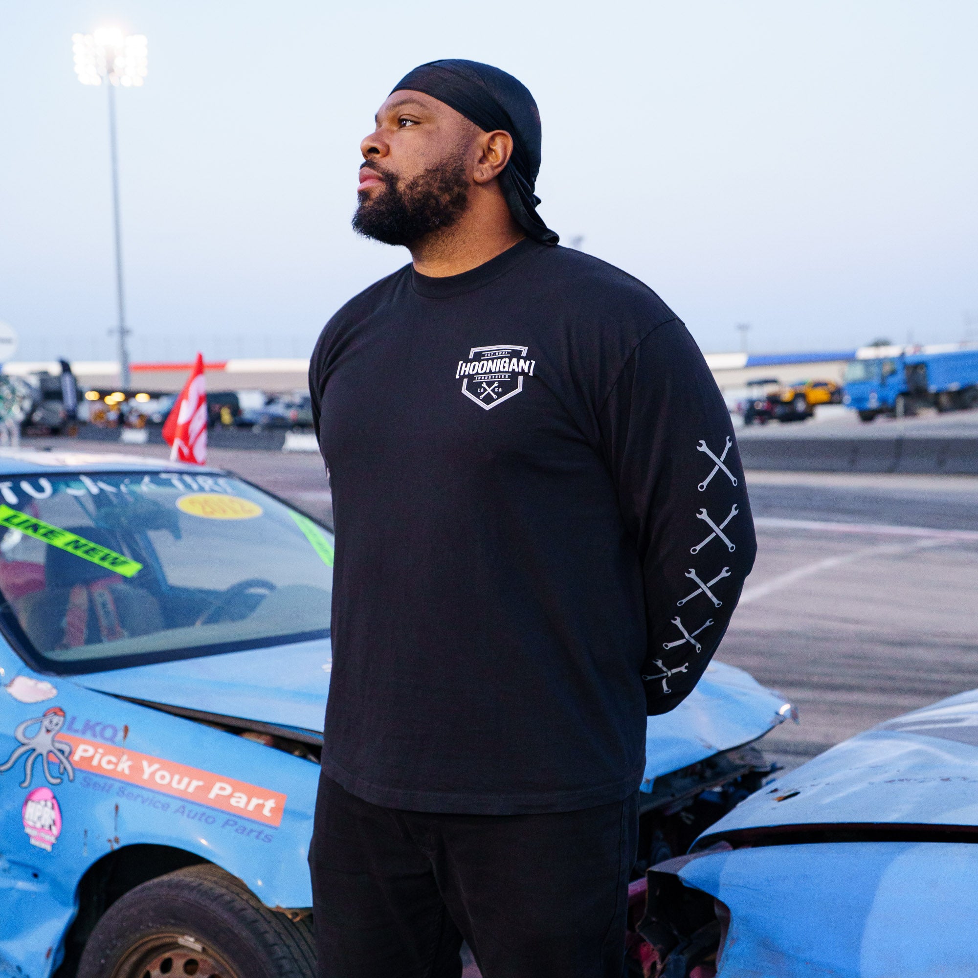Man wearing a black Hoonigan BRACKET X LOGO long sleeve T-shirt, standing beside a damaged car at a motorsport event.