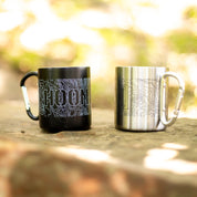 Two metal mugs with carabiner handles featuring topographic map designs and the word "HOONIGAN" on a wooden surface outdoors.