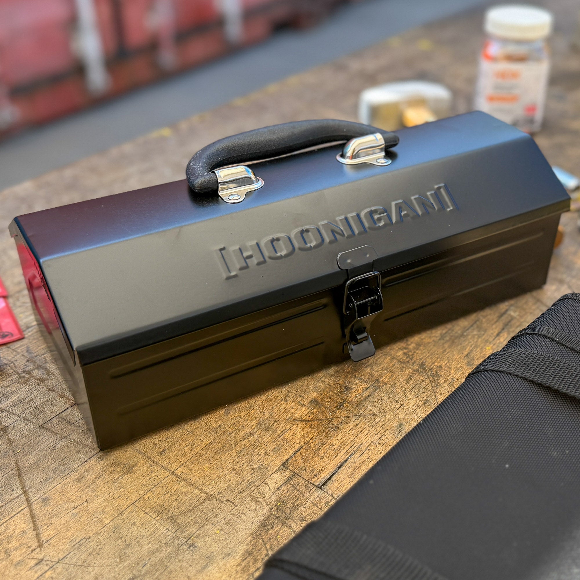 Hoonigan metal tool box with a sleek black finish and a sturdy handle, resting on a wooden surface.