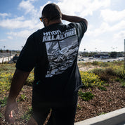 Man wearing Hoonigan WANGAN C1 Short Sleeve Tee featuring a graphic design on the back, outdoors with a scenic background.