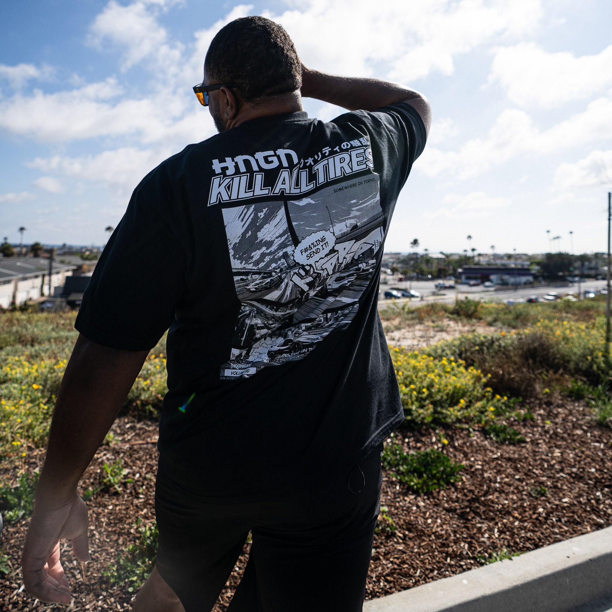 Man wearing Hoonigan WANGAN C1 Short Sleeve Tee featuring a graphic design on the back, outdoors with a scenic background.