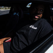 Man wearing a black Hoonigan WANGAN C1 short sleeve tee, seated in a car, looking out of the window.