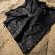 Hoonigan Oil Slick board shorts laid on sandy beach, featuring a black design with subtle logo detailing.