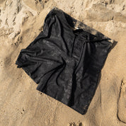 Hoonigan Oil Slick board shorts laid on sandy beach, featuring a sleek black design with subtle branding.