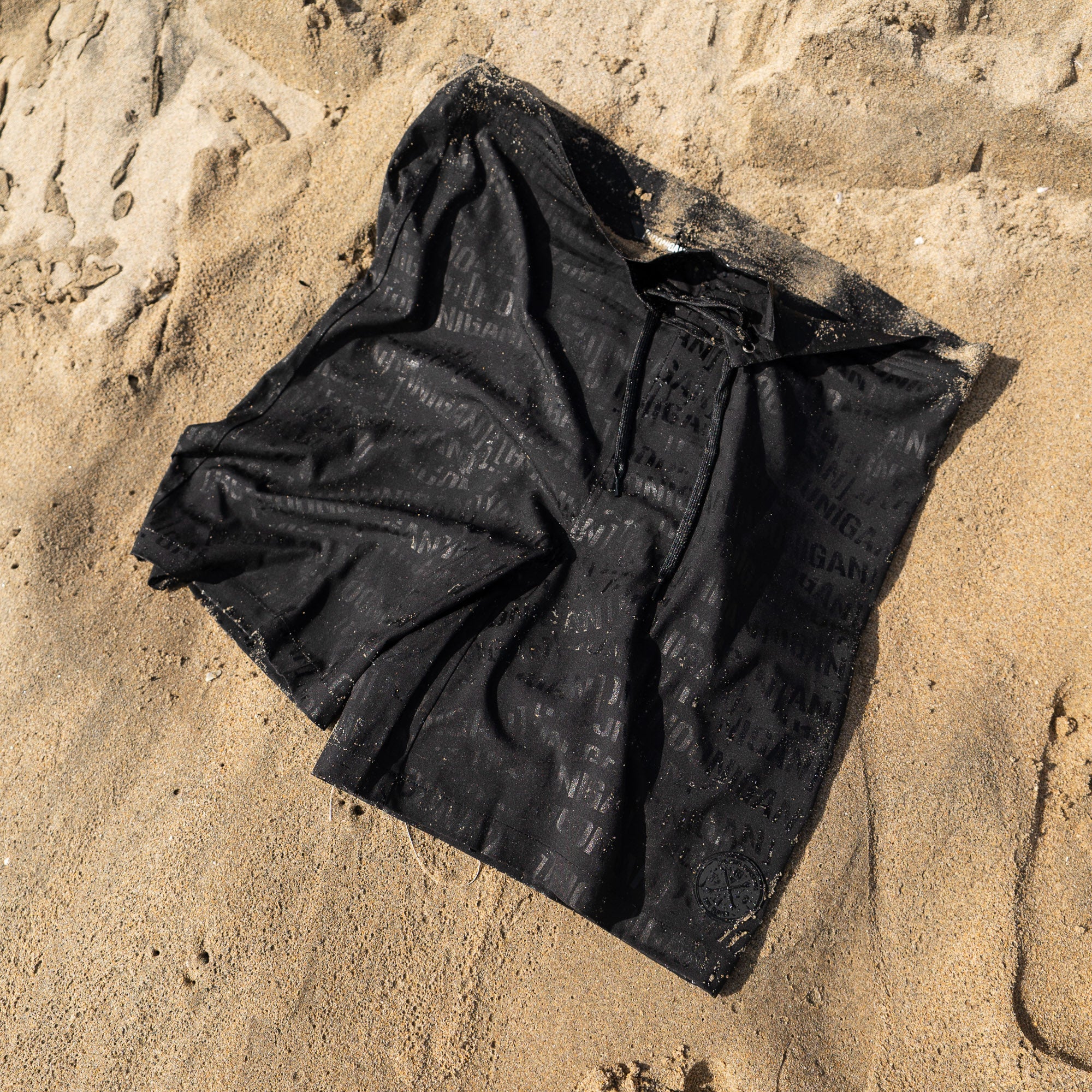 Hoonigan Oil Slick board shorts laid on sandy beach, featuring a sleek black design with subtle branding.