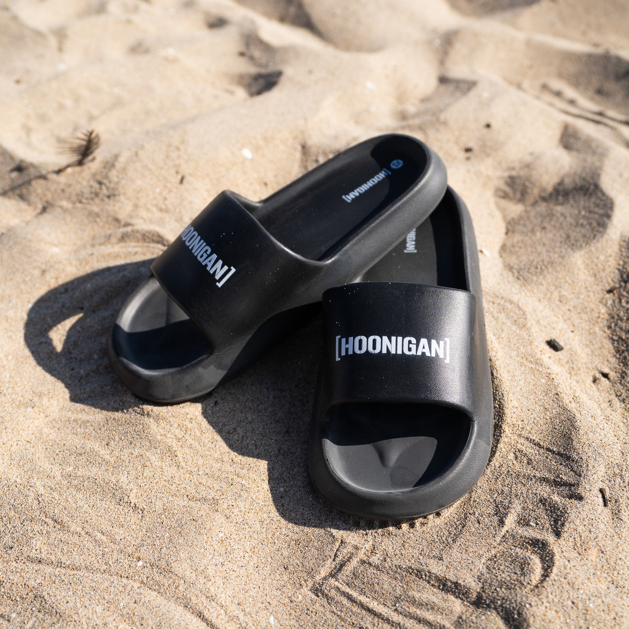 Hoonigan CENSOR BAR BRACKET LOGO slides resting on sandy beach, featuring a bold logo on a sleek black design.