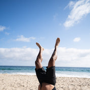 Person performing a handstand on a sandy beach, wearing Hoonigan Oil Slick board shorts against a clear blue sky.