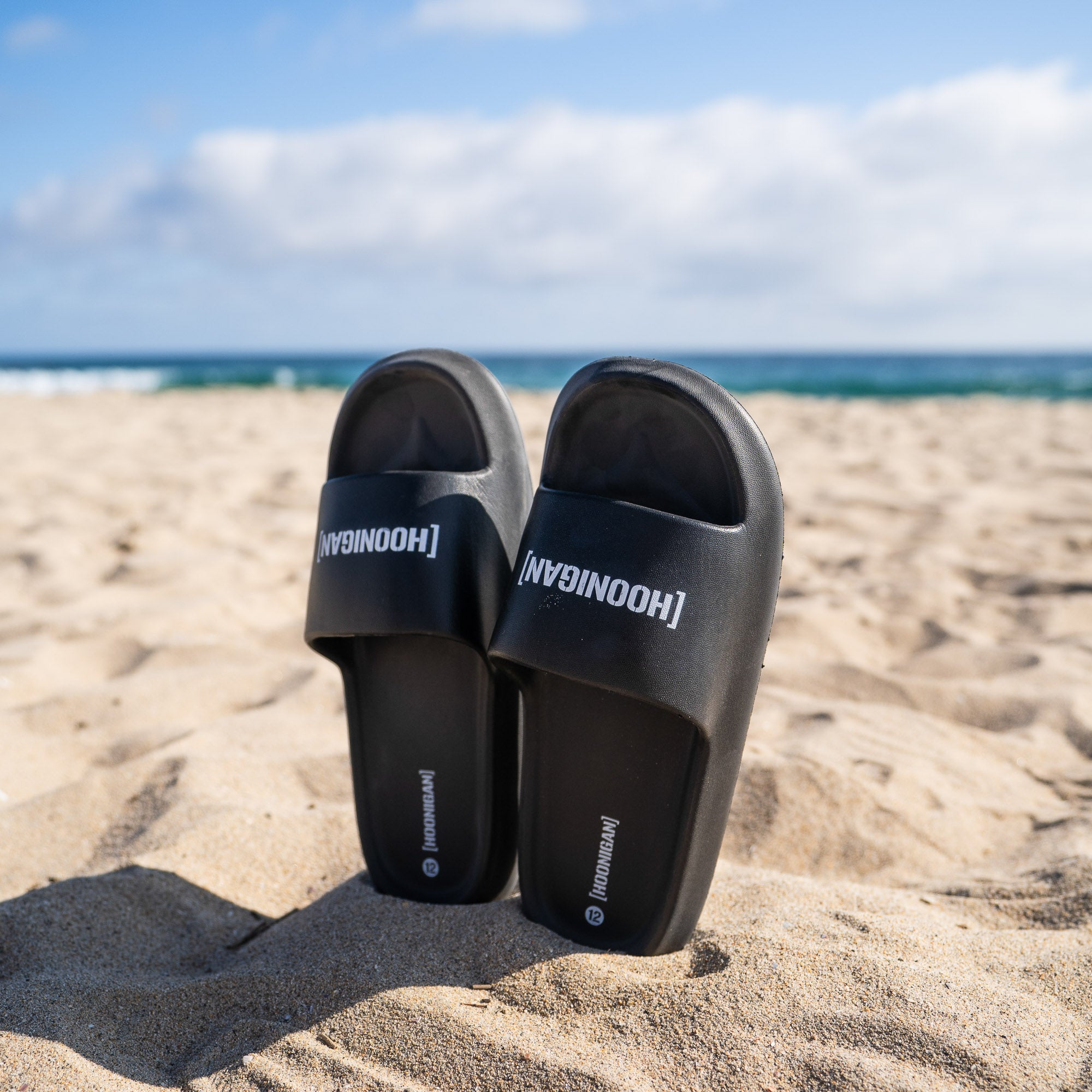 Hoonigan CENSOR BAR BRACKET LOGO slides resting on sandy beach with ocean waves in the background.