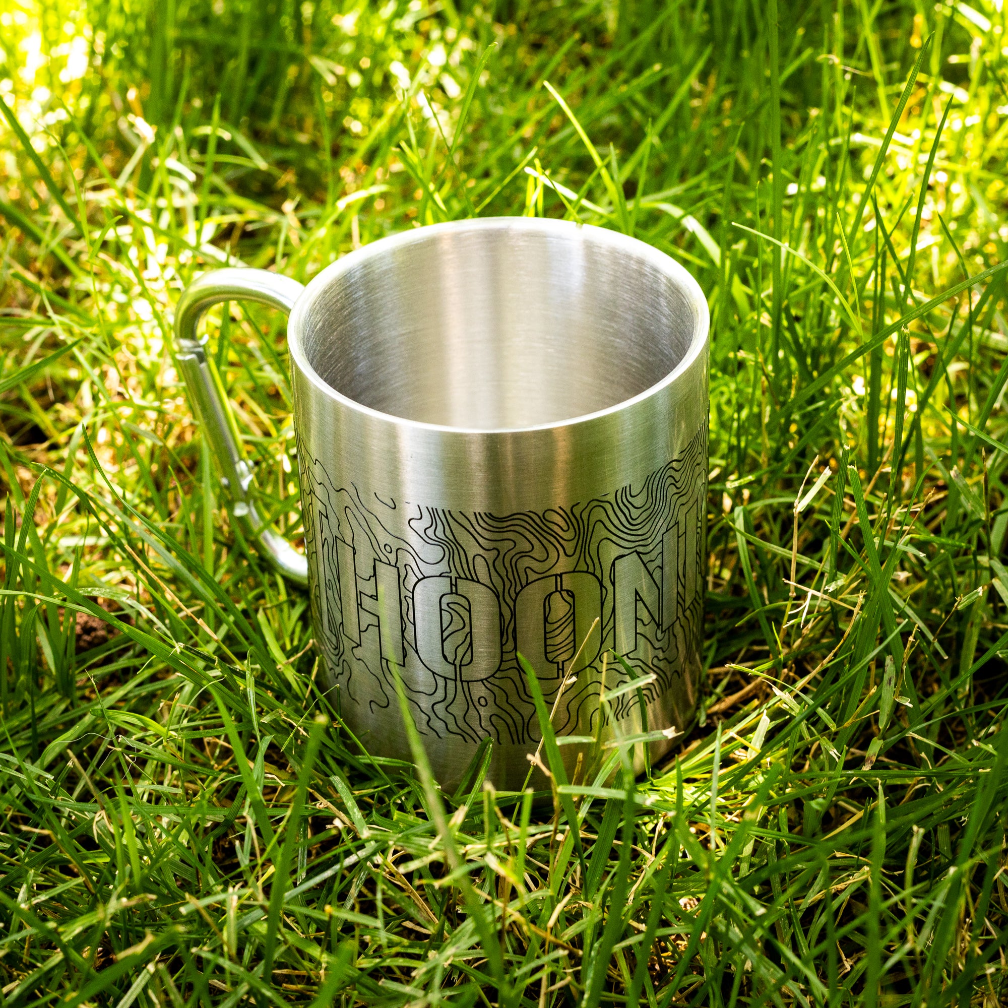 Stainless steel mug with topographic design and carabiner handle, placed outdoors on green grass.