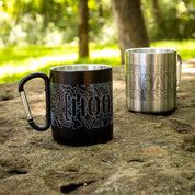 Two metal mugs with "HOONIGAN" and topographic map designs, one black with a carabiner handle, outdoors on a rock.