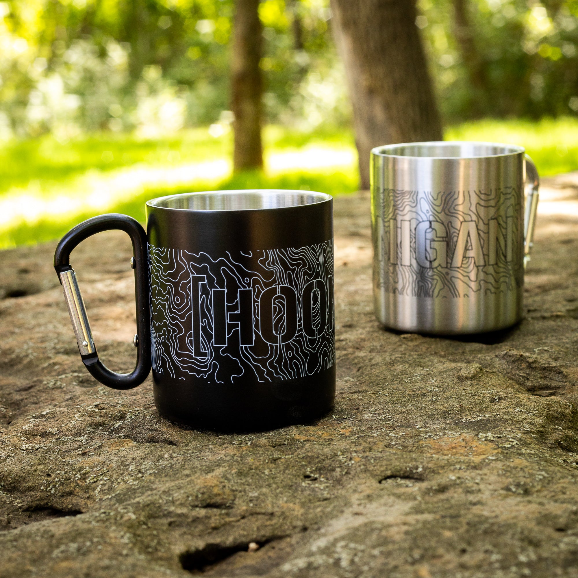 Two metal mugs with "HOONIGAN" and topographic map designs, one black with a carabiner handle, outdoors on a rock.
