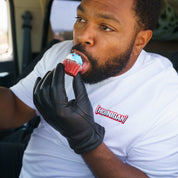 Man wearing a white Hoonigan T-shirt and black glove, licking a cupcake with blue frosting inside a vehicle.