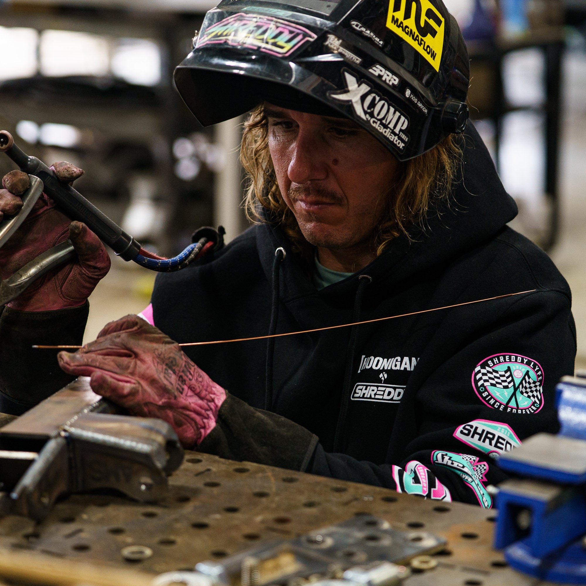 A skilled craftsman in a Hoonigan x Shreddy pullover hoodie focuses on welding in a workshop, showcasing automotive passion.