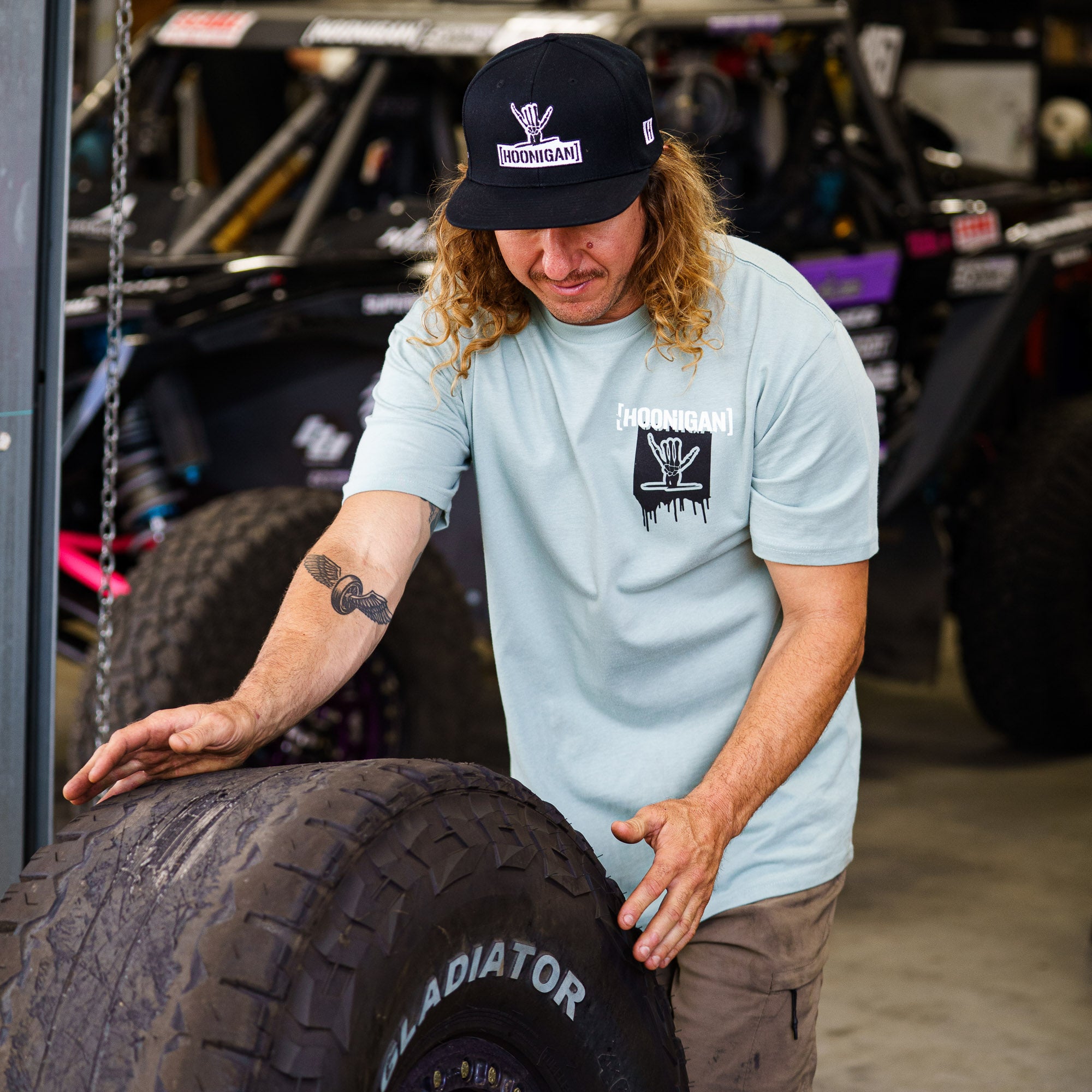 Man in a light blue short sleeve Hoonigan x Shreddy t-shirt inspects a large tire in a garage setting.