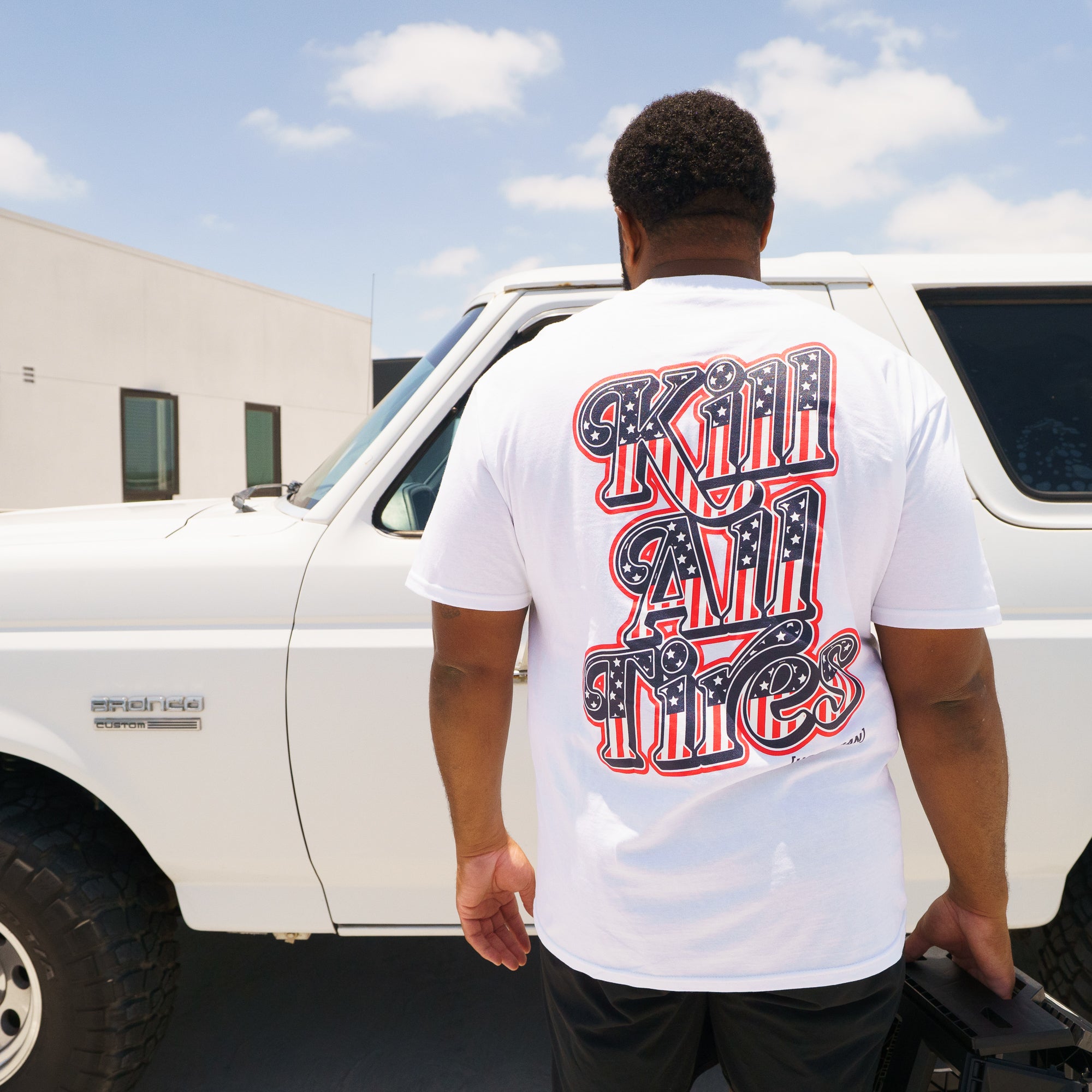 Man wearing a white T-shirt with bold red, black, and blue graphic text on the back, standing next to a white Ford Bronco.
