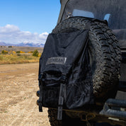 Hoonigan CENSOR BAR backpack attached to a spare tire on a rugged vehicle, ideal for outdoor adventures.