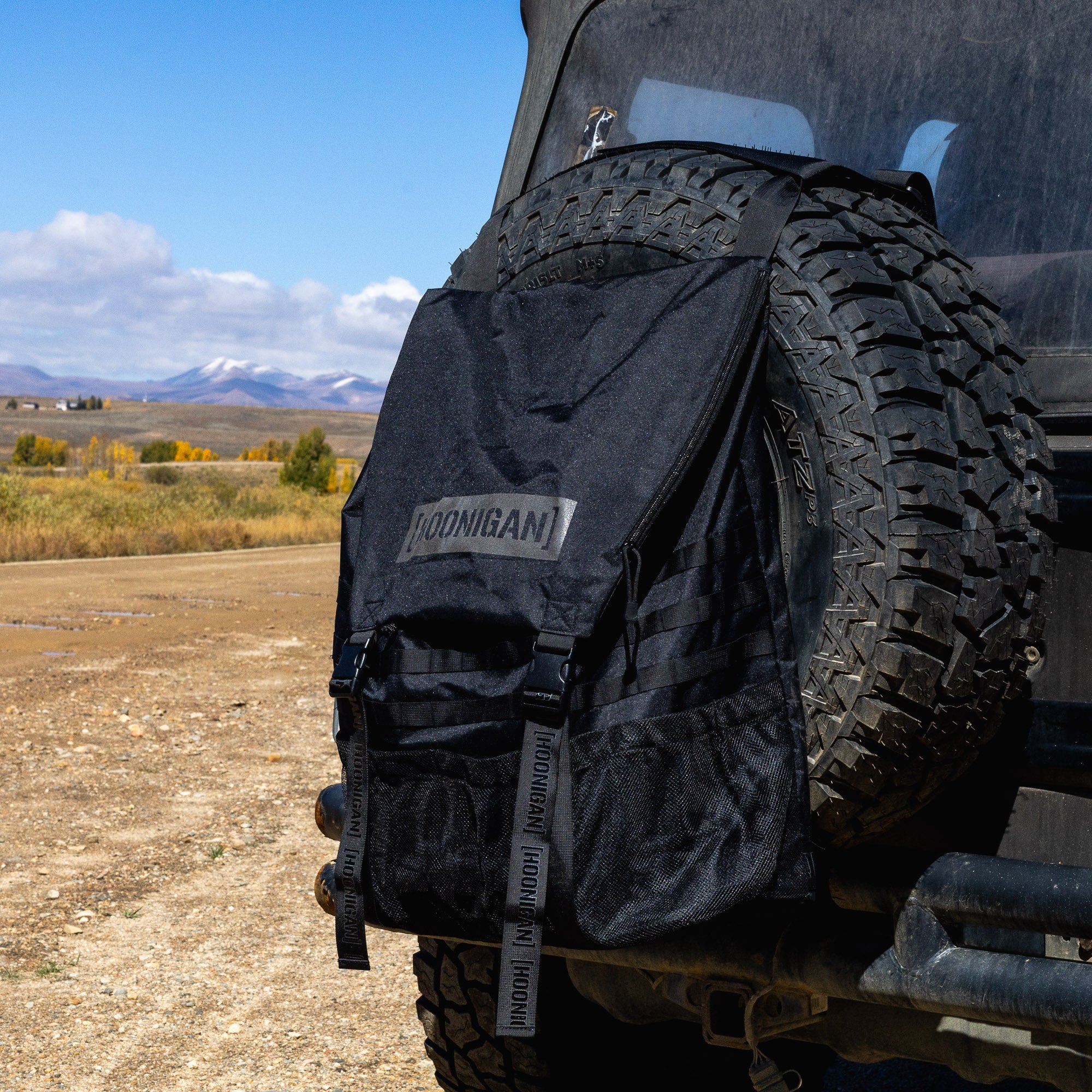 Hoonigan CENSOR BAR backpack attached to a spare tire on a rugged vehicle, ideal for outdoor adventures.