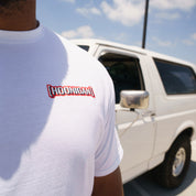 Close-up of a white T-shirt with "Hoonigan Kill All Tires" logo, worn by a person standing outdoors near a white vehicle.