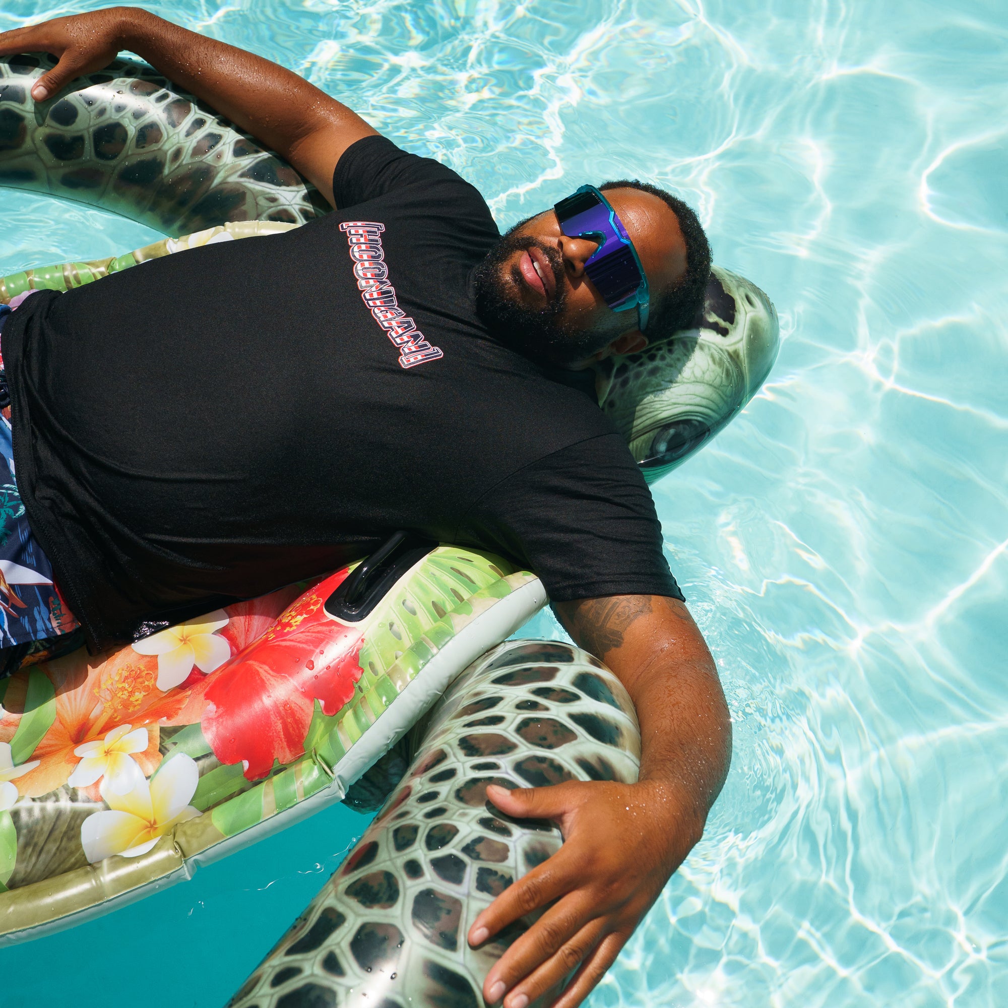 Man relaxing on a pool float wearing a black Hoonigan BRACKET USA short sleeve tee, sunglasses, and colorful swim trunks.