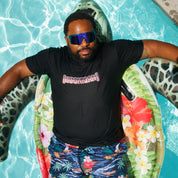 Man relaxing on a pool float wearing a black Hoonigan USA short sleeve T-shirt, tropical shorts, and sunglasses.