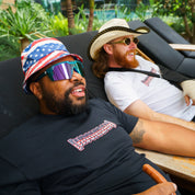 Two men relaxing outdoors, one wearing a patriotic bucket hat and sunglasses, the other with a cowboy hat and sunglasses.