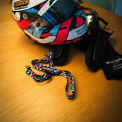 Hoonigan x Lia Block SCRIPT lanyard displayed with a colorful racing helmet and gloves on a wooden surface.