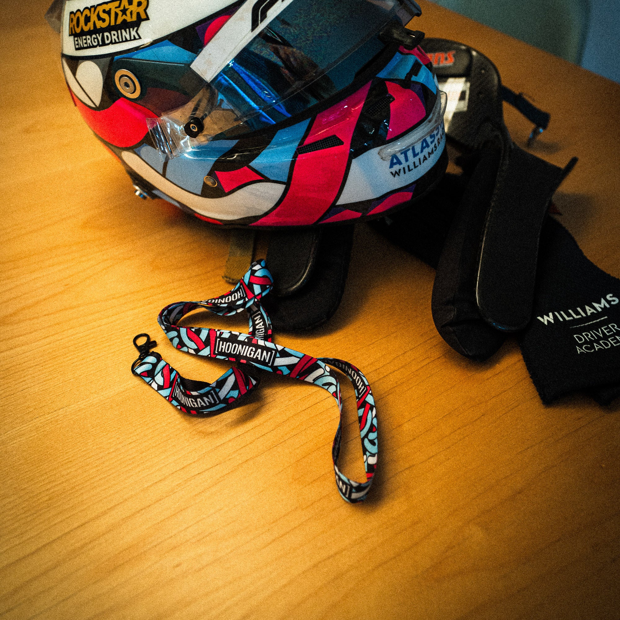 Hoonigan x Lia Block SCRIPT lanyard displayed with a colorful racing helmet and gloves on a wooden surface.