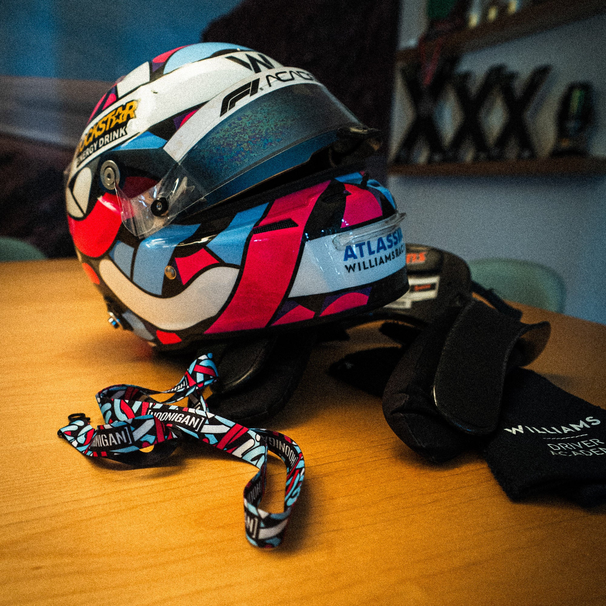 Hoonigan x Lia Block SCRIPT lanyard displayed next to a colorful racing helmet and gloves on a wooden table.