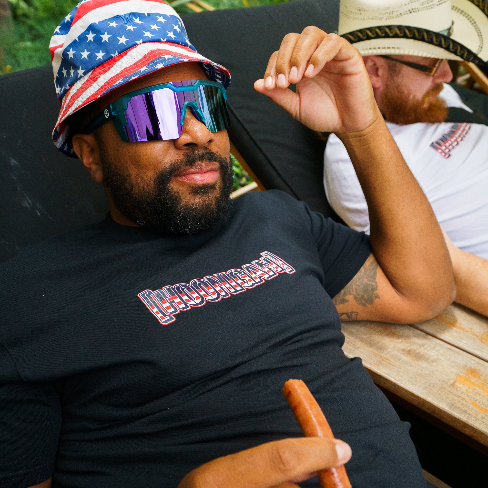 Man wearing a patriotic bucket hat and reflective sunglasses, relaxing outdoors in a black Hoonigan T-shirt.