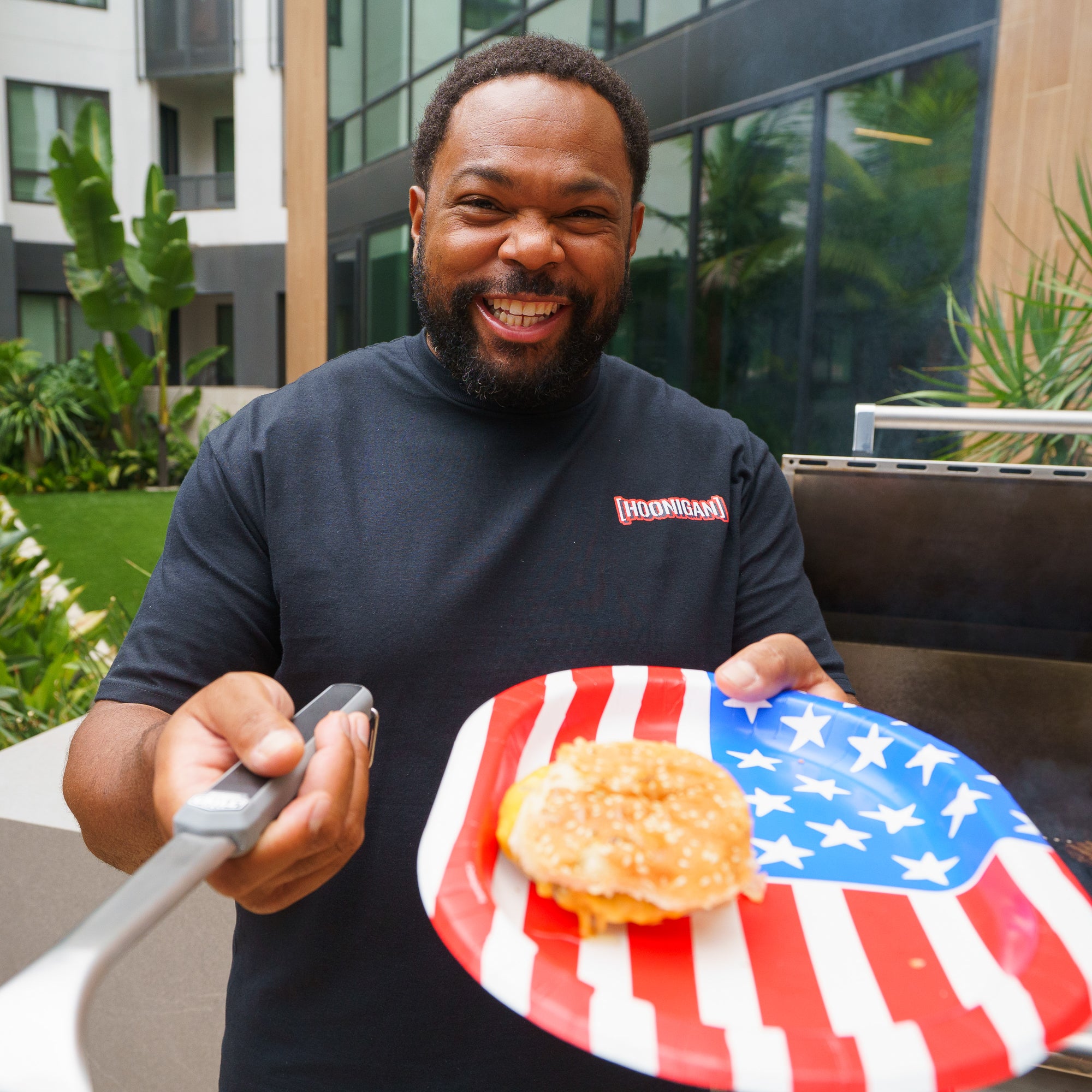 Man smiling and holding a plate with a burger on a patriotic American flag-themed tray, outdoors in a modern residential area.