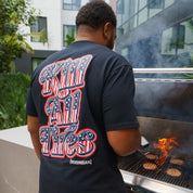 Back view of a man wearing a black Hoonigan Kill All Tires USA short sleeve T-shirt with bold patriotic graphic text, grilling burgers outdoors.