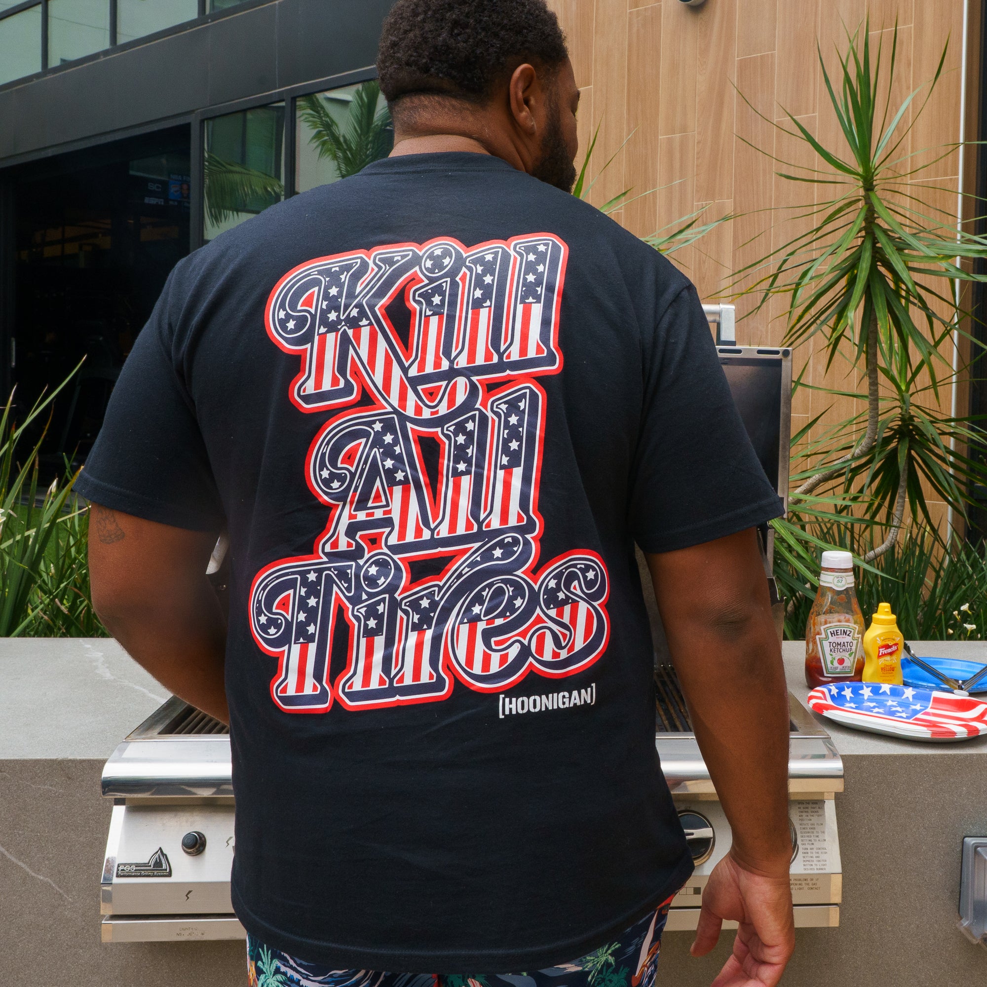Back view of a man wearing a black Hoonigan Kill All Tires short sleeve T-shirt with bold red, white, and blue graphic text.