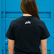 Back view of a woman wearing a black Hoonigan x Lia Block CENSOR BAR short sleeve t-shirt against a blue background.