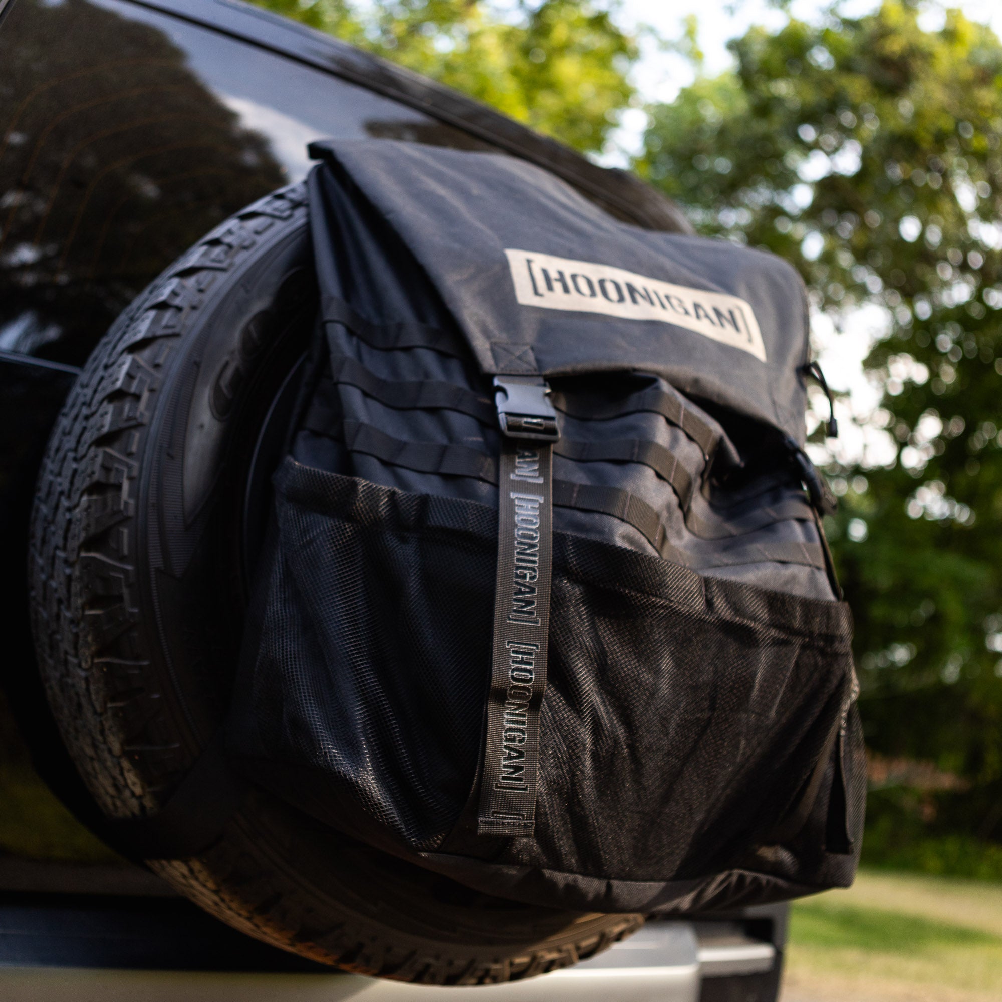 Hoonigan CENSOR BAR backpack attached to a spare tire, featuring a sleek black design and durable straps.