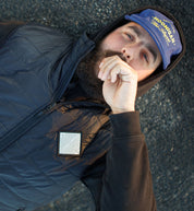 Man lying on asphalt wearing a black quilted puffer vest with a reflective patch, blue Hooinghan cap, and black hoodie.