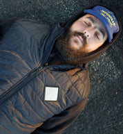 Man lying on ground wearing a black quilted puffer vest, hoodie, and a blue cap with "HONGAN" logo.